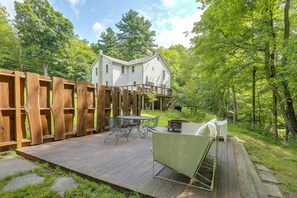 A wooden deck with outdoor furniture, a white house in the background, and a forested area. - Hot Tub, Sauna, Fire Pit! Catskills Family Retreat (Ellenville)