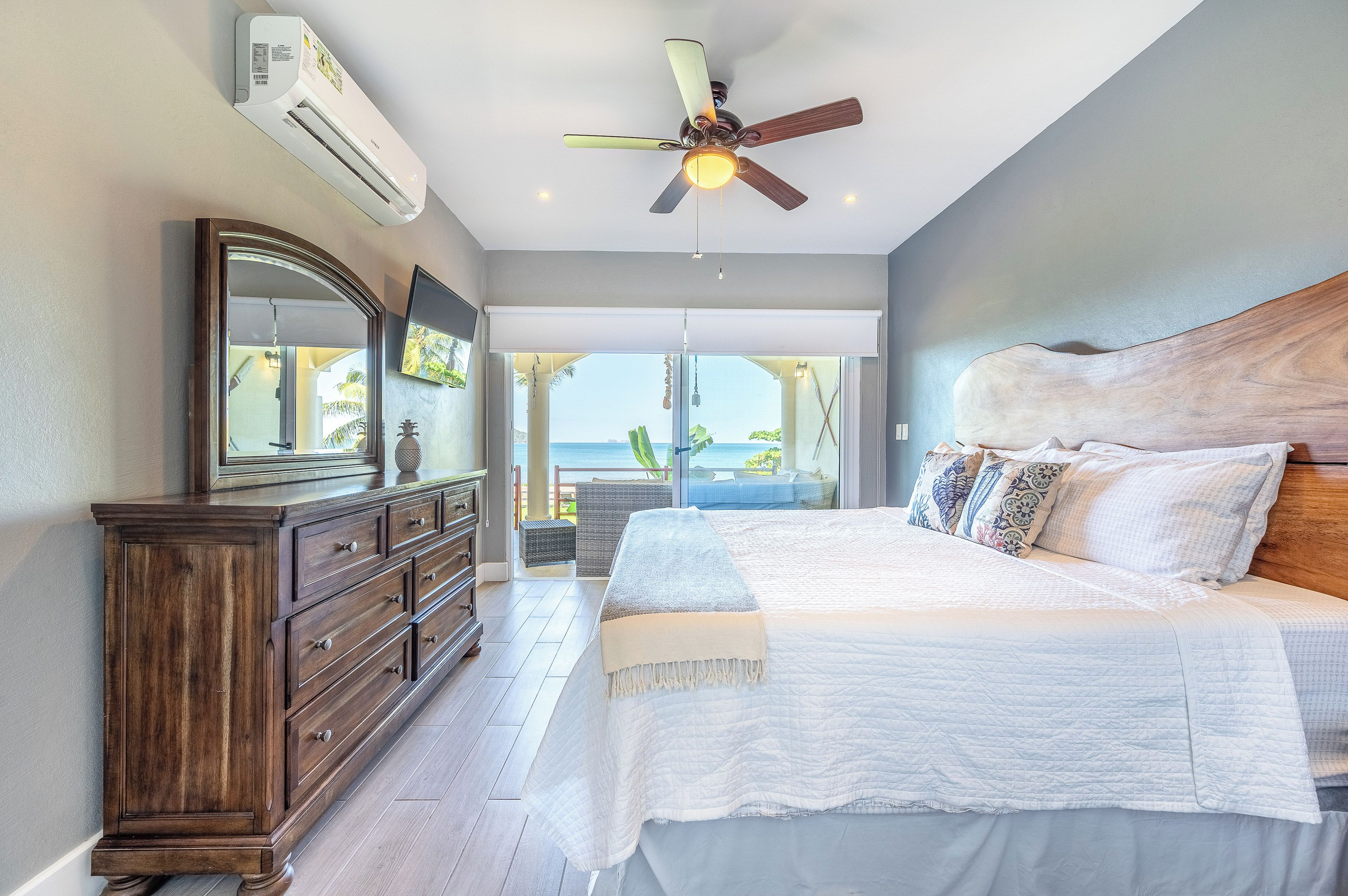 A bedroom with a large bed, a wooden dresser, a ceiling fan, and a view of the ocean through a sliding door.