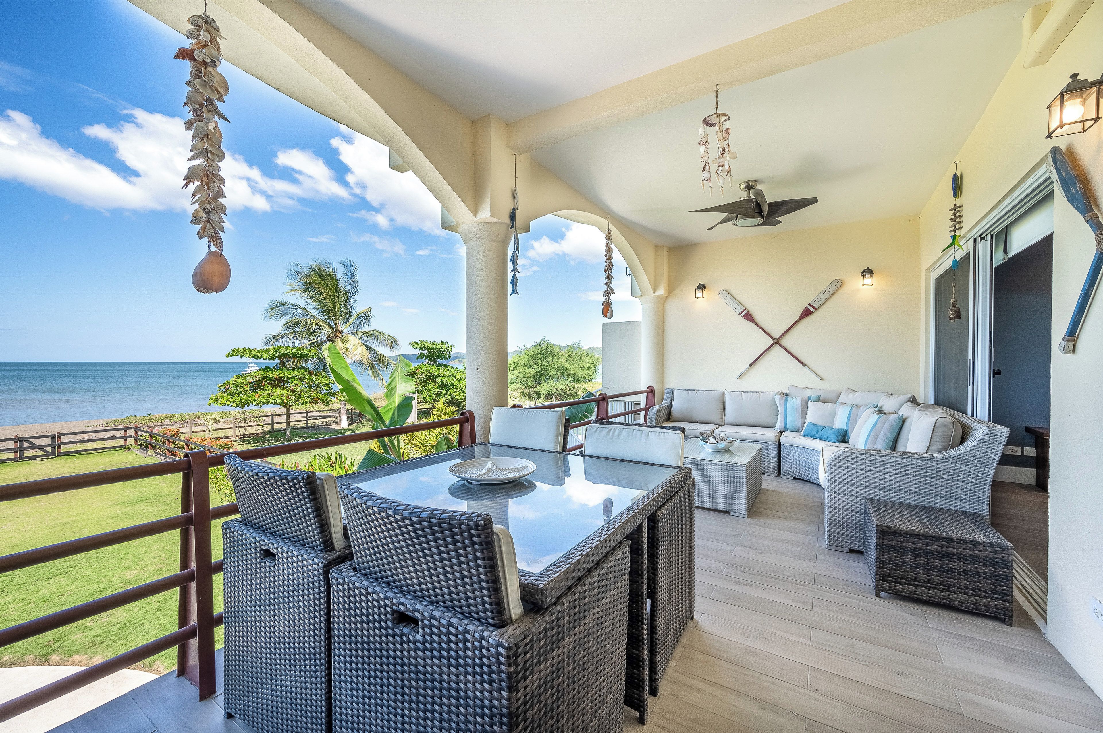 A covered outdoor seating area with wicker furniture, a glass table, and a view of the ocean.