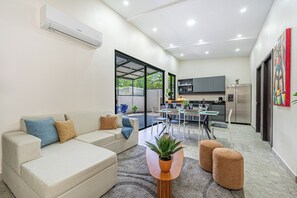 Un salon moderne avec un canapé, des poufs et une table basse. On y trouve également un coin repas avec une table et des chaises, ainsi qu'une cuisine équipée d'un réfrigérateur et de placards.