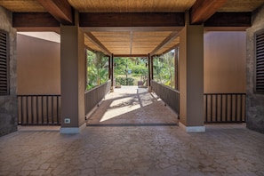 Uma galeria coberta com piso de pedra, grades de metal e vista para o verde.