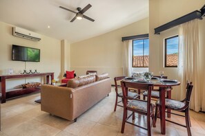 A living room with a sofa, a dining table set for four, a flat-screen TV, and a ceiling fan. - Sunny 2br: Balcony, Pool, Kitchen, Walk to Beach (Sardinal)