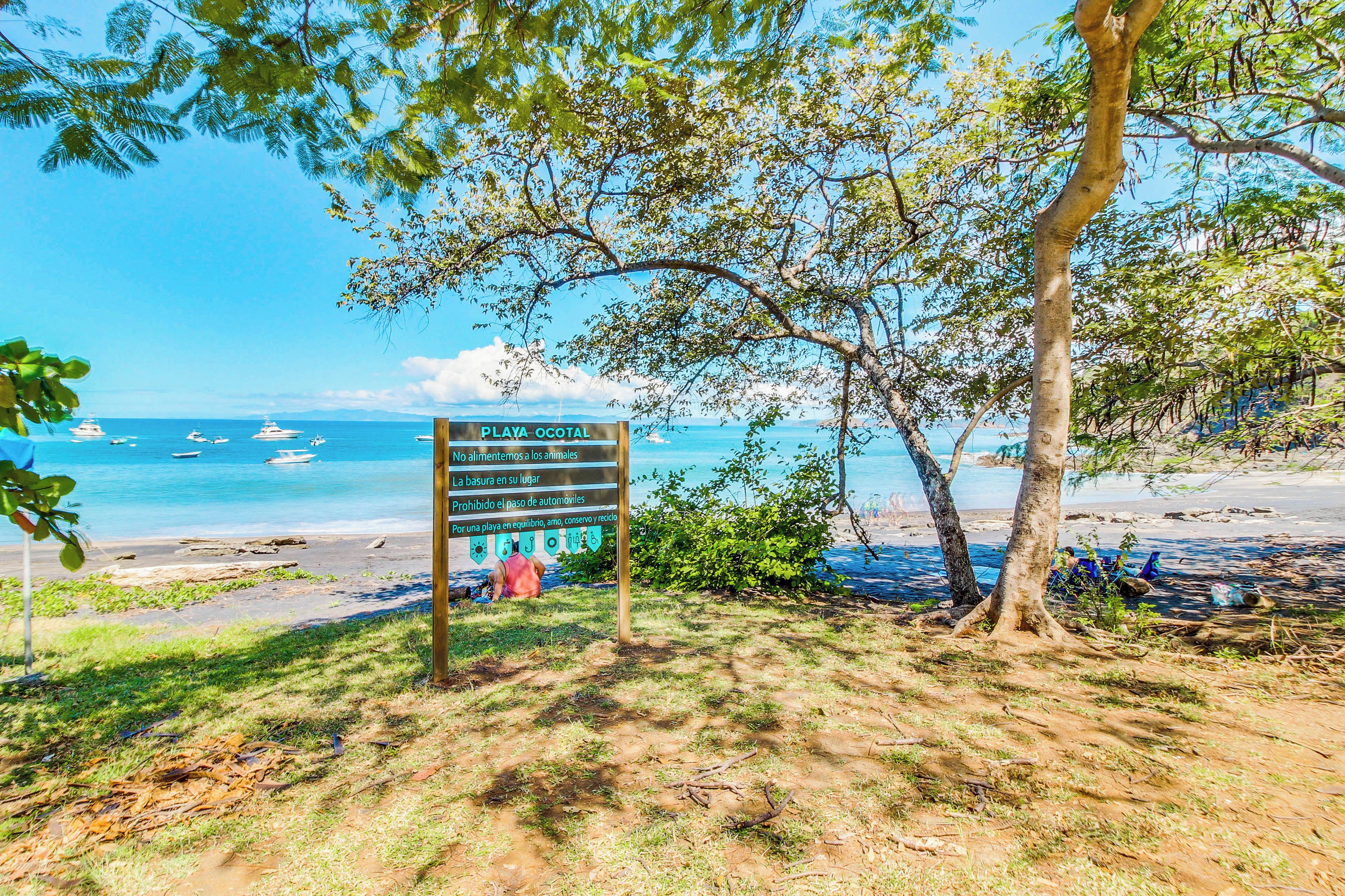 A beach sign with environmental guidelines and a view of the ocean with boats.
