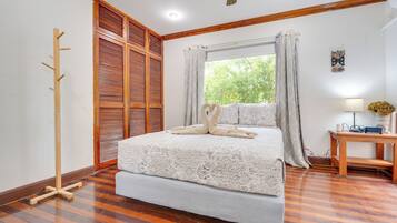 A bedroom with a large bed, wooden floor, a wooden coat rack, a window with curtains, and a bedside table with a lamp.