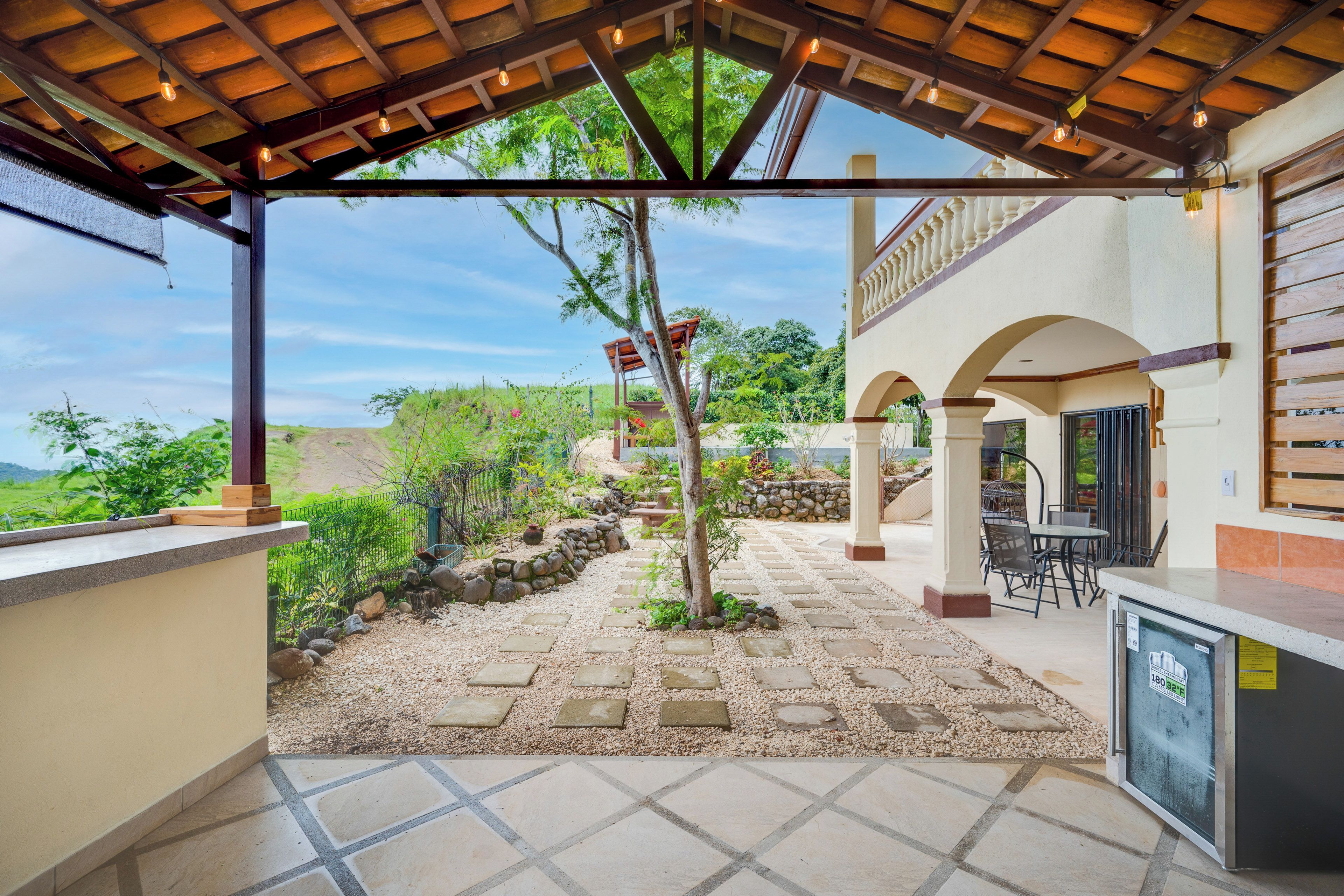 A covered outdoor area with a stone pathway, a pergola with hanging lights, and a view of the surrounding landscape.