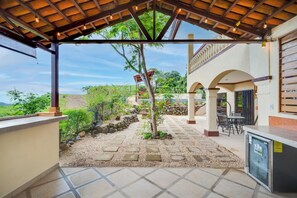 A covered outdoor area with a stone pathway, a pergola with hanging lights, and a view of the surrounding landscape.