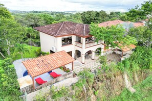 Una casa de dos pisos con un balcón, un patio con sillas reclinables rojas y vistas a la vegetación circundante.