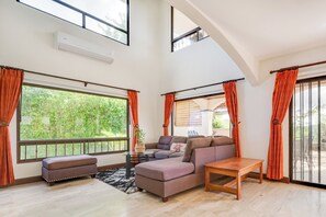A spacious living area with a large sofa, ottoman, and a wooden coffee table. There are multiple windows with orange curtains, and a view of greenery outside. - Private Pool, Mtn-view! 3BR Playas Coco Home (Sardinal)