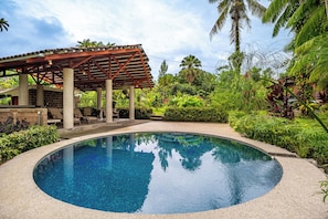 A clear blue pool surrounded by lush greenery and a covered seating area.
