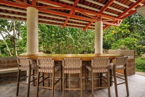 An outdoor dining area with a round wooden table and six woven chairs, surrounded by lush greenery and a wooden pergola structure.
