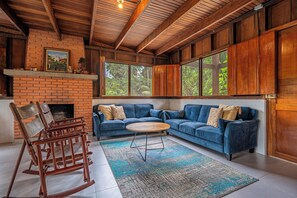 A cozy living room with a brick fireplace, blue sofa, wooden rocking chairs, and large windows.