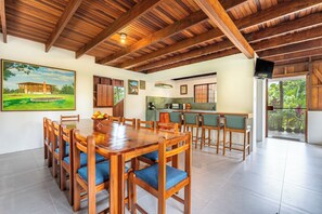 A spacious dining area with a wooden table and chairs, a kitchen in the background, and a large window with a view of greenery.