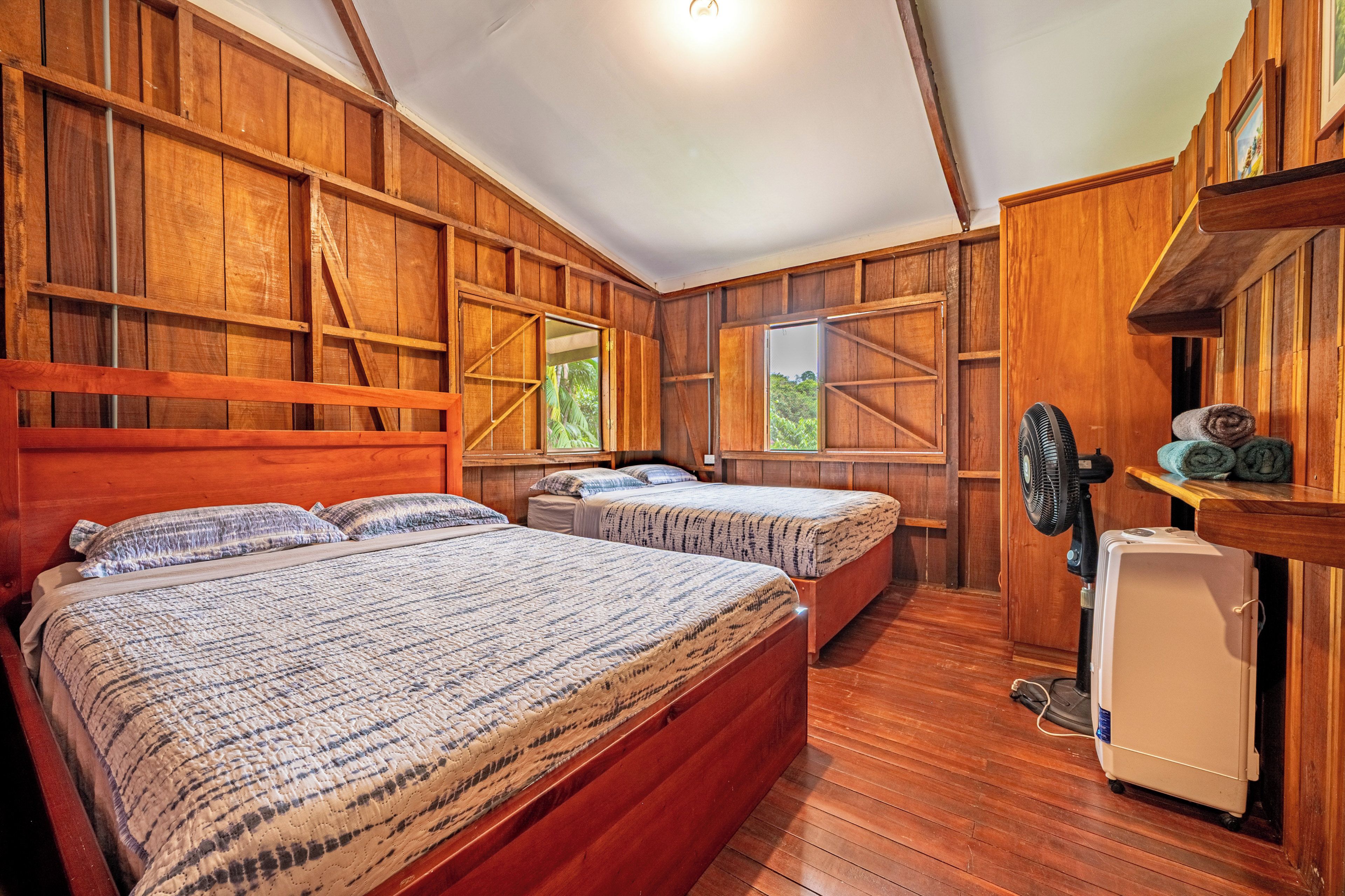A room with two beds, wooden walls, a fan, and a small refrigerator.