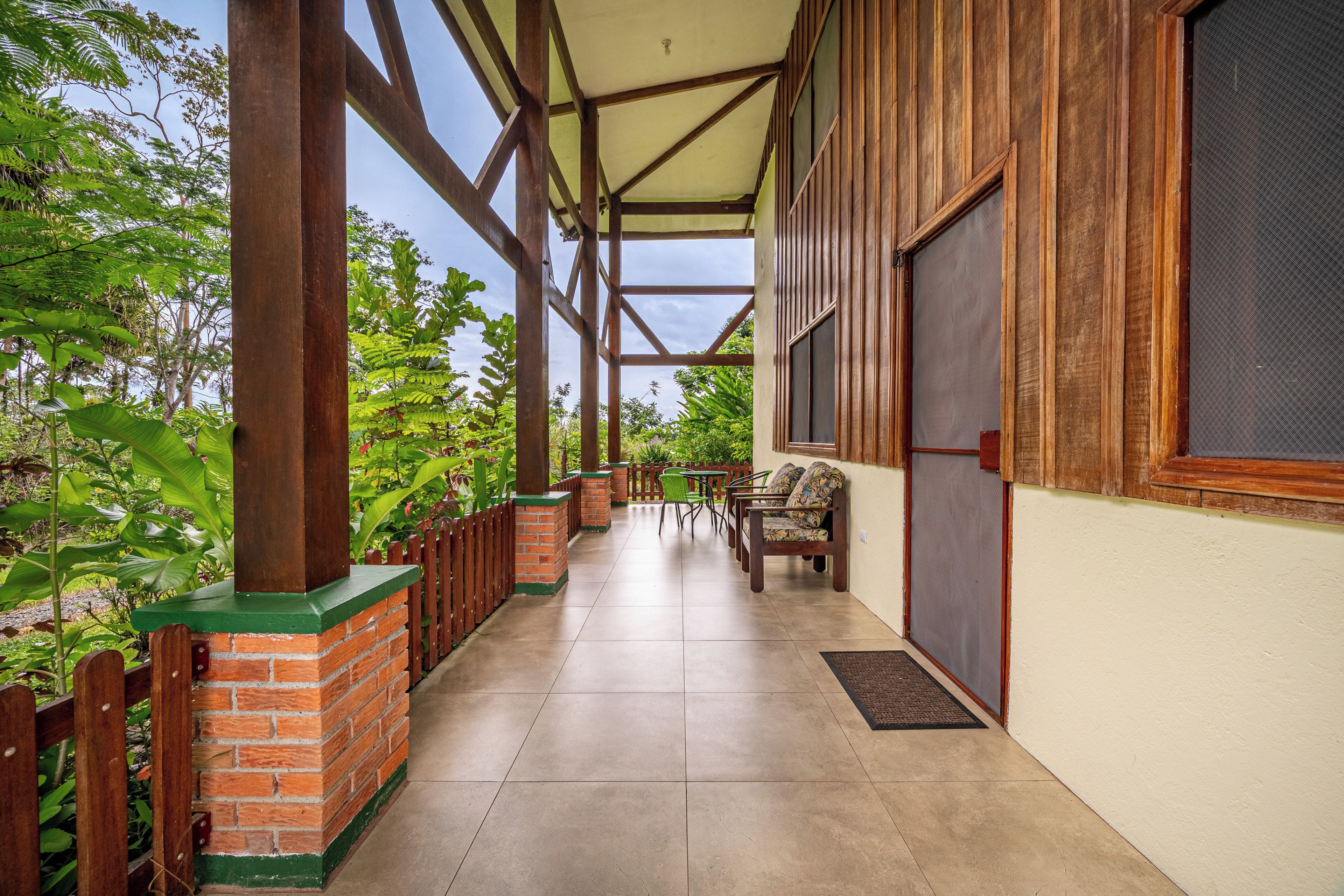 A covered outdoor area with a brick column, wooden beams, a tiled floor, and a seating area with chairs.
