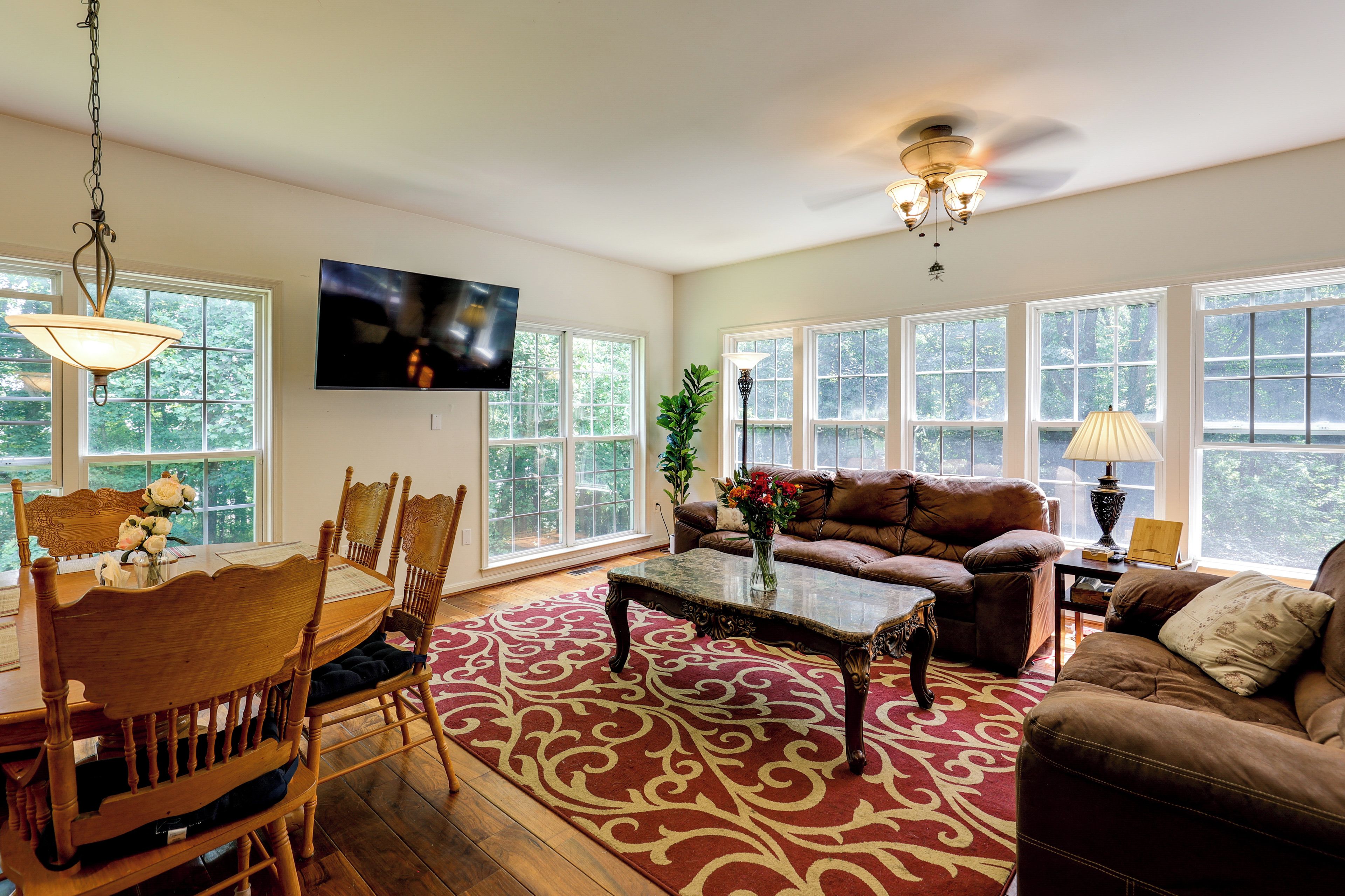 A living room with a dining area, a television mounted on the wall, and large windows.