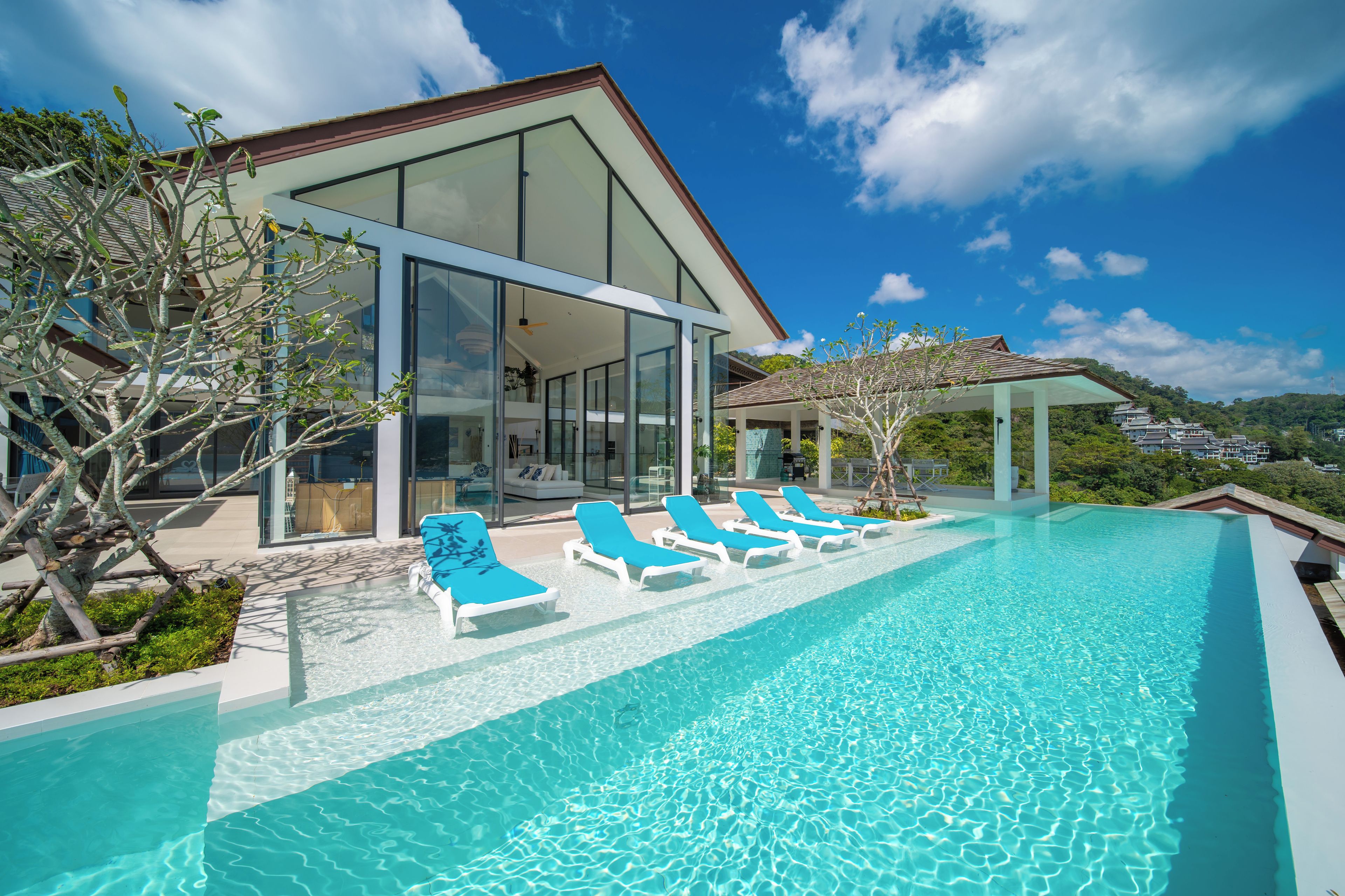 A modern house with a glass facade, a swimming pool with lounge chairs, and a view of the surrounding landscape.