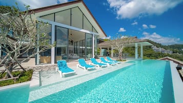 A modern house with a glass facade, a swimming pool with lounge chairs, and a view of the surrounding landscape.