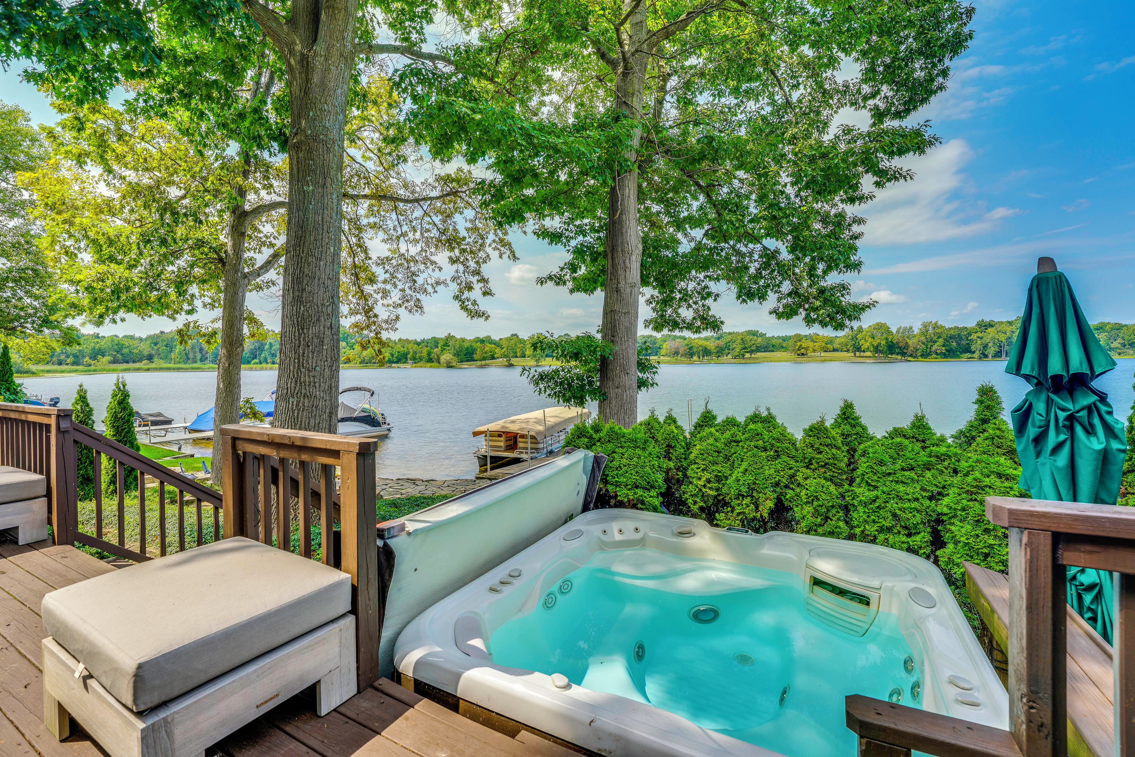 A hot tub on a wooden deck with a lake view, surrounded by trees and a boat dock.