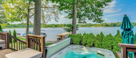 A hot tub on a wooden deck with a lake view, surrounded by trees and a boat dock.