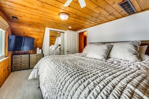 Une chambre avec un plafond en bois, un lit recouvert d’une couverture matelassée, un téléviseur posé sur une commode en bois, et une porte donnant sur une autre pièce.