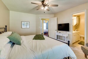 Une chambre à coucher avec un lit, un ventilateur de plafond, une télévision et une salle de bain visible à travers une porte ouverte.
