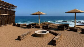 A beach with two wooden umbrellas, a fire pit, and a thatched structure.