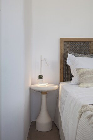 A corner of a room with a white wall, a bedside table with a white lamp, a small potted plant, and a bed with white bedding and a textured headboard. - Villa Akrotini with Sea view and Hot Tub (Kokkino Chorio)