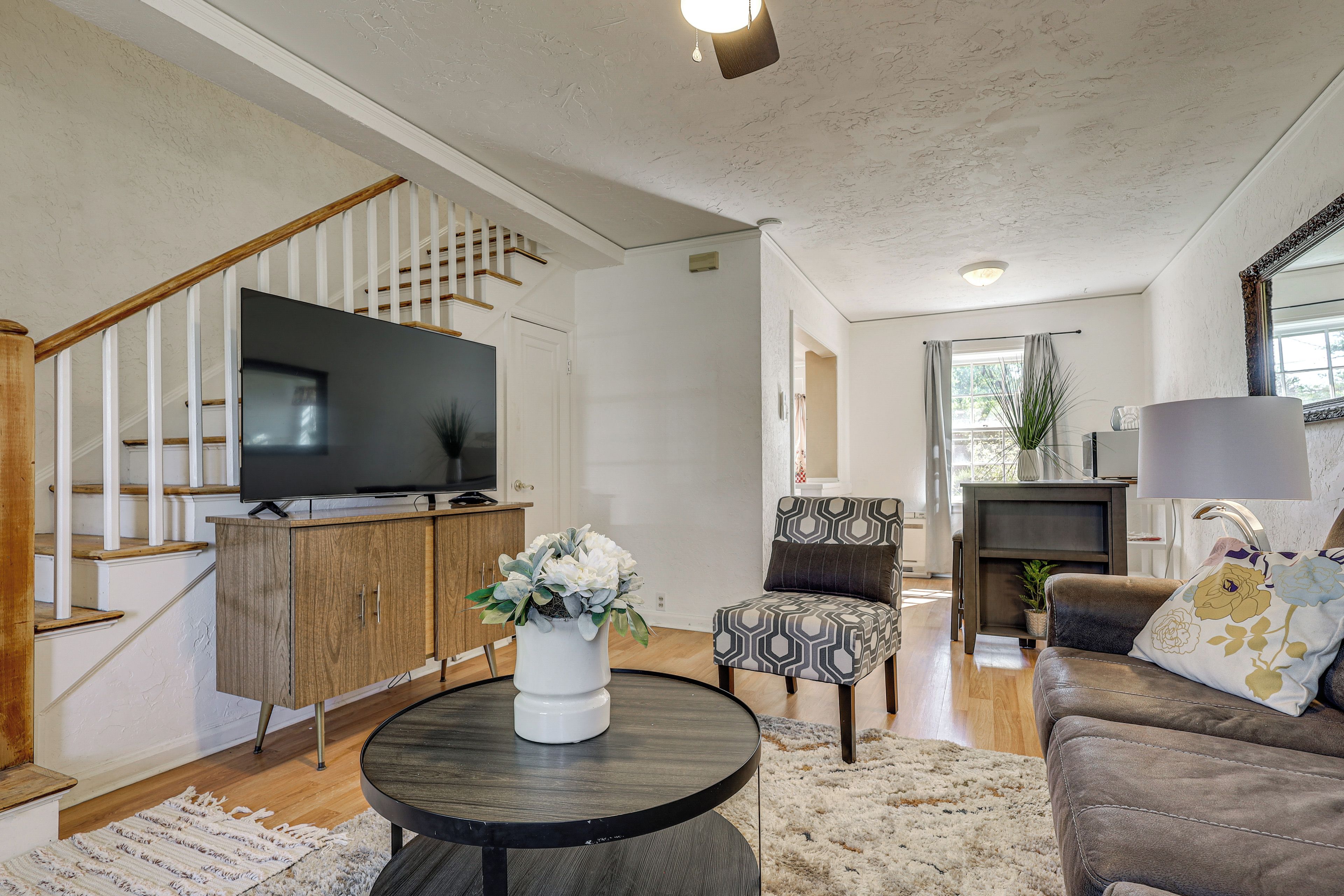 A living room with a staircase, a flat-screen TV, a round coffee table, a patterned armchair, and a sofa.
