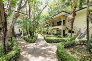 A pathway flanked by neatly trimmed hedges and trees, leading to a building with a balcony and a staircase.