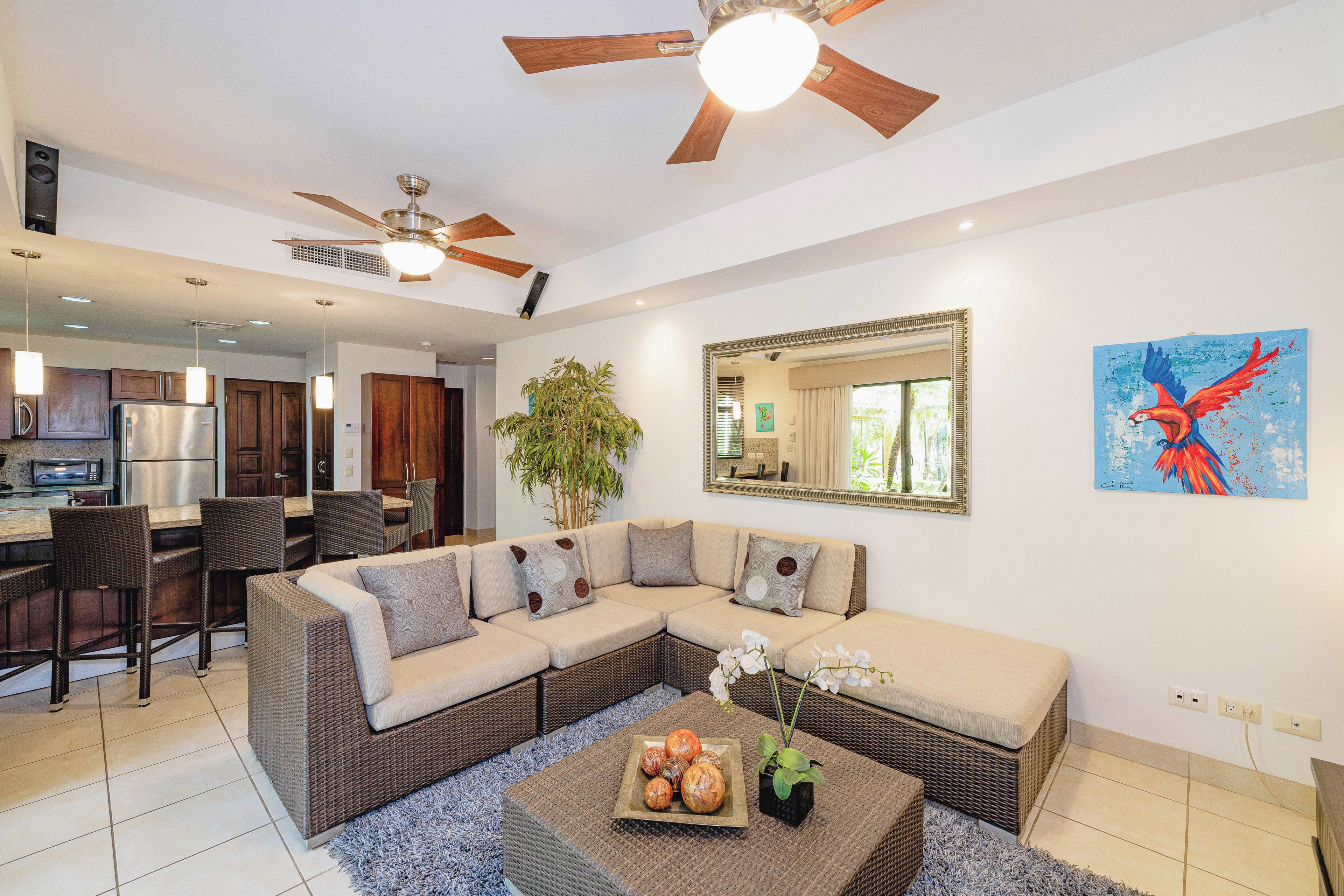 Uma sala de estar moderna com sofá seccional, uma área de jantar com cadeiras de vime, uma cozinha com eletrodomésticos de aço inoxidável e um ventilador de teto.