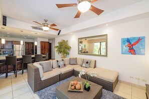 A modern living room with a sectional sofa, a dining area with wicker chairs, a kitchen with stainless steel appliances, and a ceiling fan.