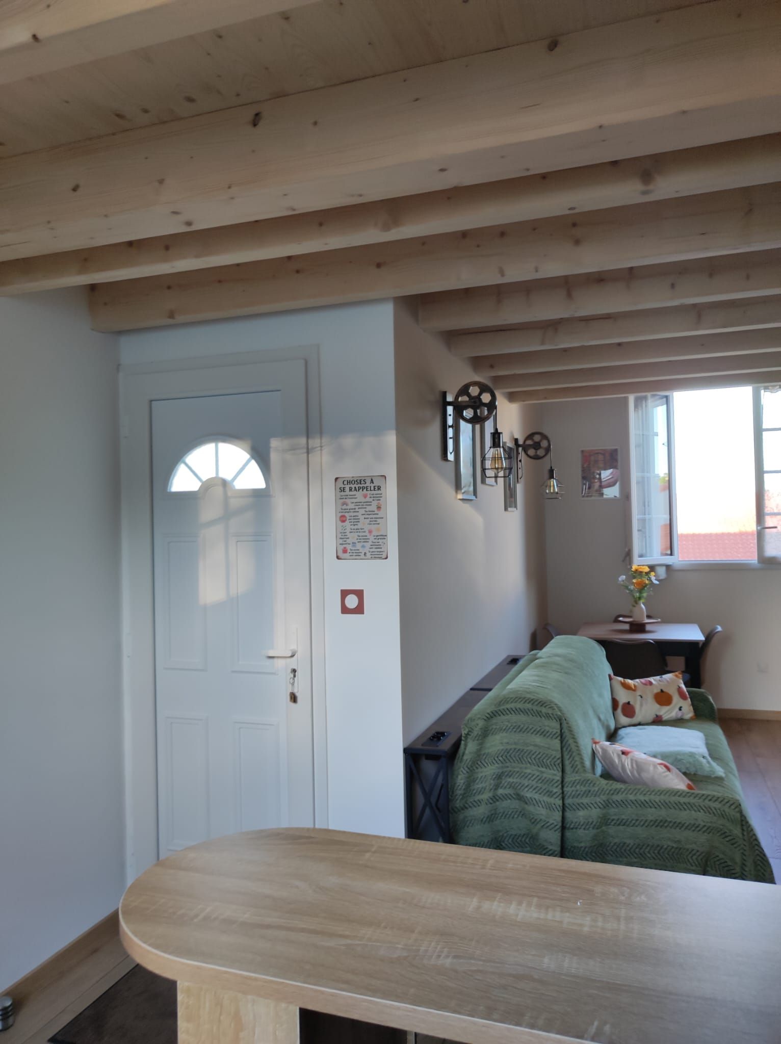 Una habitación con techo de madera, una puerta blanca, un sofá verde y una pequeña mesa con un jarrón de flores.
