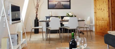 Un salon moderne comprenant un coin repas, un téléviseur fixé sur un meuble blanc, une porte en bois et une table en verre avec une bouteille et un verre.