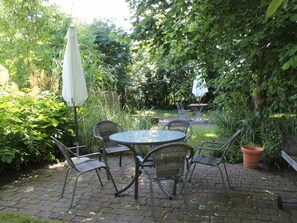 Eine Terrasse im Freien mit einem runden Tisch und vier Stühlen, umgeben von Pflanzen und einem weißen Sonnenschirm.