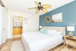 A bedroom with a bed, bedside tables, a ceiling fan, a wall-mounted shelf with plants, and a painting. - Waikiki Banyan 25th-Floor-Ocean View 1BR with Parking (Honolulu)