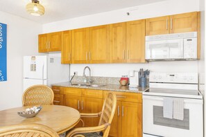 A kitchen with wooden cabinets, a microwave, a stove, and a dining area with a table and chairs. - Waikiki Banyan 25th-Floor-Ocean View 1BR with Parking (Honolulu)