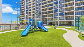Un terrain de jeux avec des toboggans et un espace piscine, entouré d’un bâtiment doté de nombreux balcons, sous un ciel dégagé.