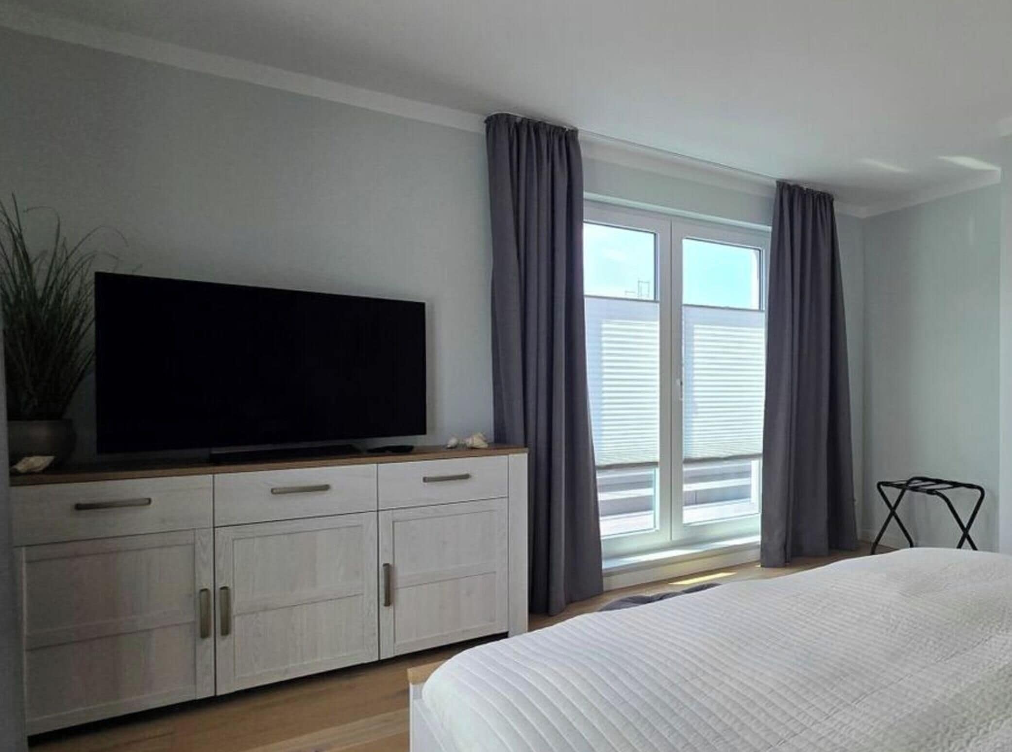 Un dormitorio con una cama, un televisor sobre un mueble, una ventana con cortinas y una planta.