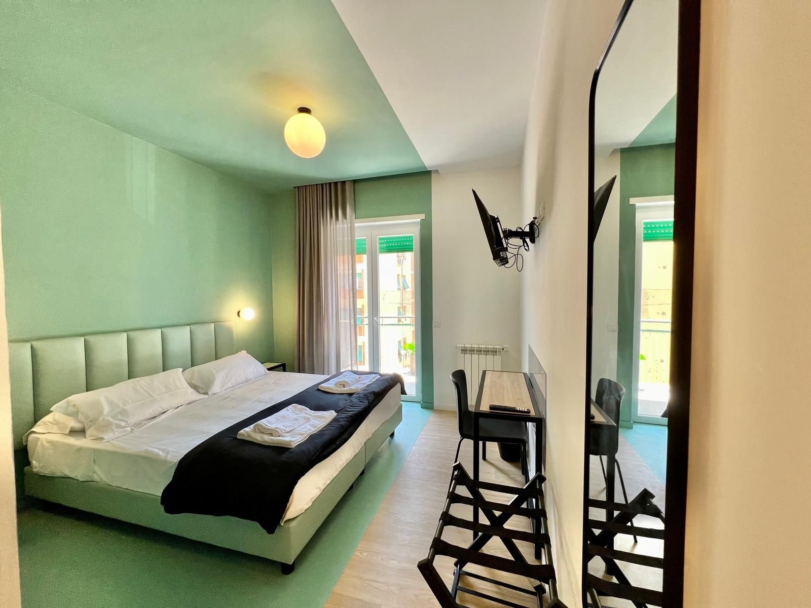 Une chambre d’hôtel avec un lit, un bureau et une chaise. Il y a une fenêtre avec des rideaux, un radiateur et un luminaire fixé au mur.