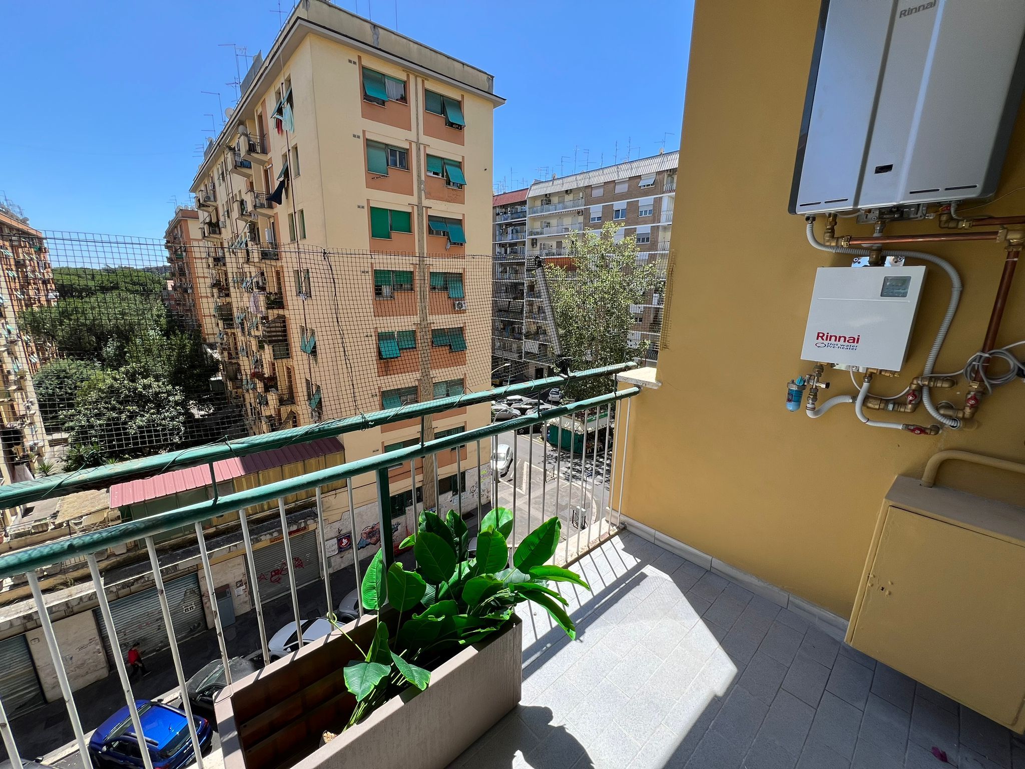 A balcony with a potted plant, a water heater, and a view of a residential building.