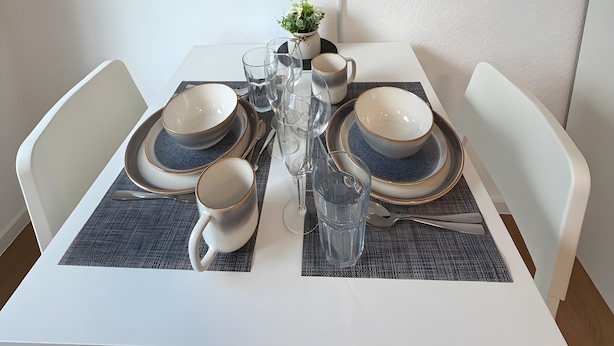 A dining table set with plates, bowls, glasses, and cutlery, with a flat-screen TV mounted above.