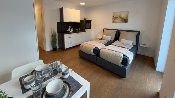Um apartamento moderno com cozinha compacta, área de refeições com mesa posta para quatro pessoas e uma cama grande com cabeceira.
