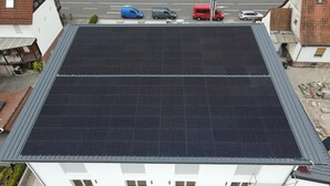 A building with a flat roof covered in solar panels, a paved driveway, and a street with parked cars.