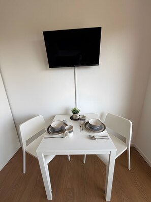 Ein Essbereich mit einem weißen Tisch, zwei Stühlen, einem an der Wand montierten Flachbildfernseher und einer kleinen Pflanze auf dem Tisch.
