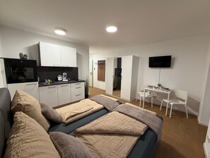 Eine moderne Wohnung mit einer Küchenzeile, einem Bett mit beigefarbener Bettwäsche, einem Schreibtisch mit Stuhl und einem wandmontierten Flachbildfernseher.
