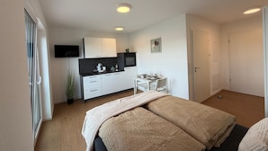 Quarto moderno com cama grande, mesa de cabeceira e uma pequena cozinha no fundo.