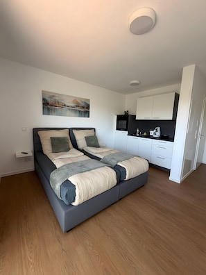 A modern bedroom with a large bed, a small bedside table, a painting on the wall, and a kitchenette in the corner.