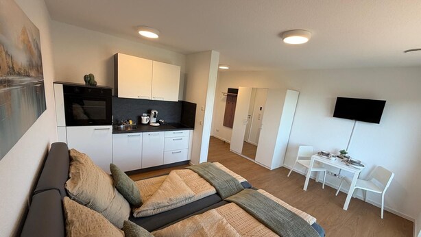 Quarto de hotel moderno com cama, cozinha compacta, área de refeições e televisão.