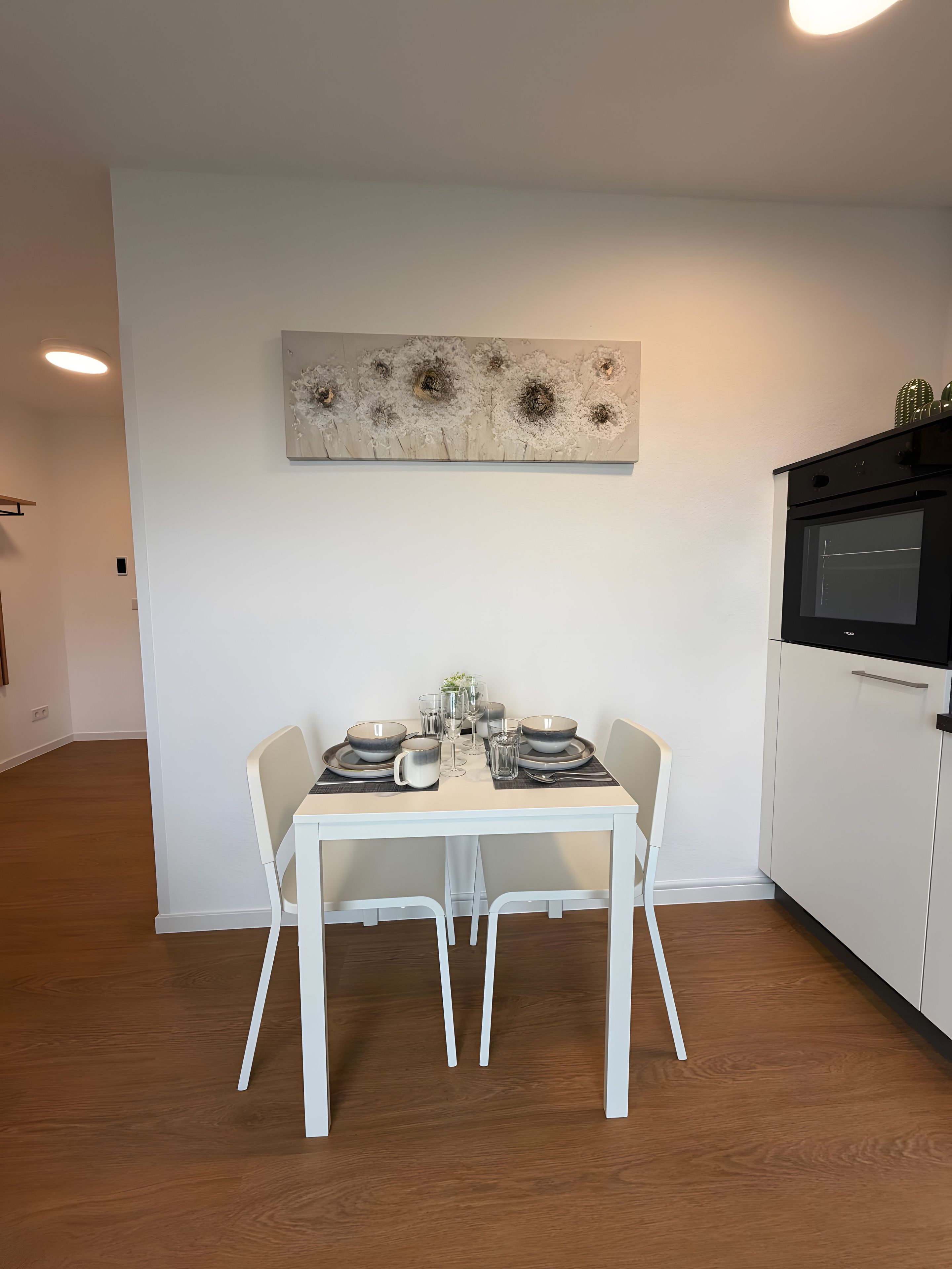 Ein Essbereich mit einem weißen Tisch, gedeckt für zwei Personen, eine moderne Küche mit einem schwarzen Backofen und einem dekorativen Wandbild über dem Tisch.