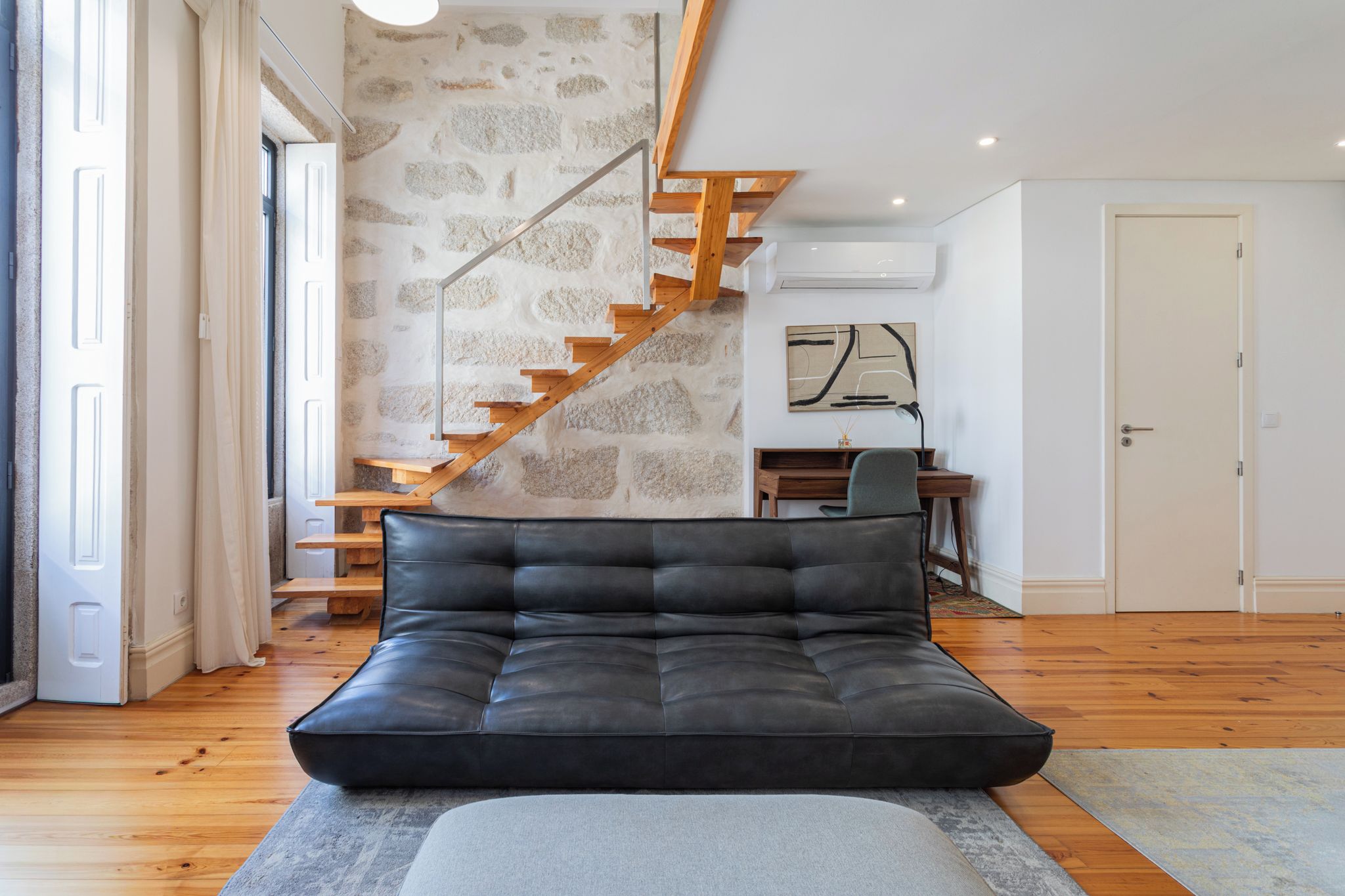 Uma sala de estar moderna com um sofá de couro preto, uma escada de madeira e uma mesa de escritório com uma cadeira.